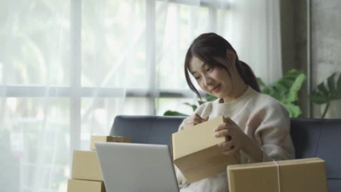 亚洲妇女坐在沙发上，坐在家里的盒子和笔记本电脑从客户网上业务中小企业销售概念