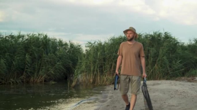 夏天日落时分，白种大胡子男子手持登陆网和鱼竿，在乡间的河边钓鱼。