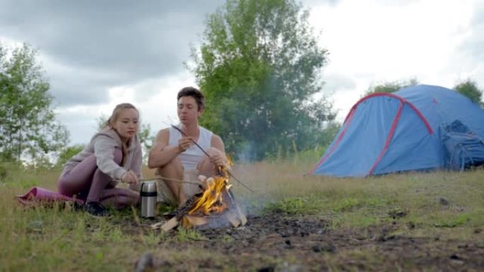 Young couple sits by campfire frying and eating ma