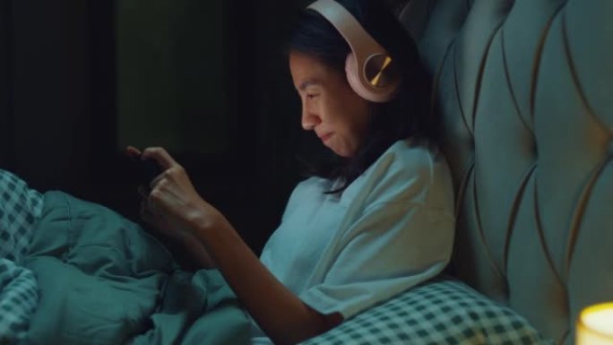 年轻的亚洲女孩媒体成瘾在床上戴着耳机笑在流媒体在线视频点播中观看最喜欢的电影，晚上在家点播视频。