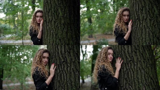 快乐的年轻美女看着相机拥抱户外绿色公园的树干。平静自信的高加索女士的肖像摆姿势在户外享受大自然的美丽