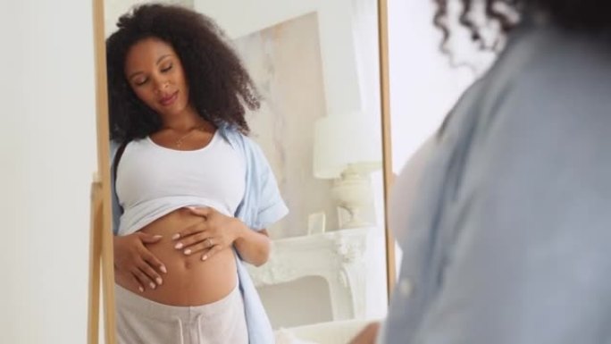 黑人孕妇早上在镜子里欣赏她的肚子。享受积极的情绪。黑人年轻美丽的未来母亲在家里快乐地抚摸着她的肚子。