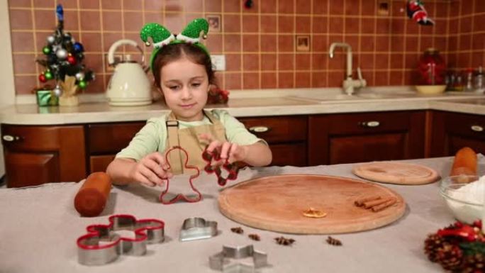 可爱的小女孩穿着精灵箍和厨师围裙，与站在厨房桌子上的姜饼切刀一起玩