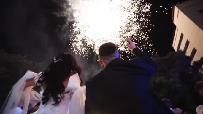 新郎和新娘在天空观看烟花。恋爱中的情侣在婚礼结束时接吻。兴奋的客人看着火展。