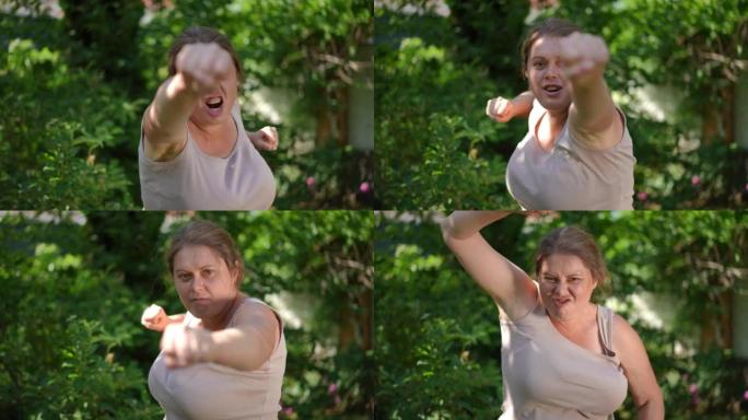 愤怒的大码女人在慢动作中看着相机拳击阴影的前视图肖像。有动力的高加索超重女运动员训练在夏季花园户外摆