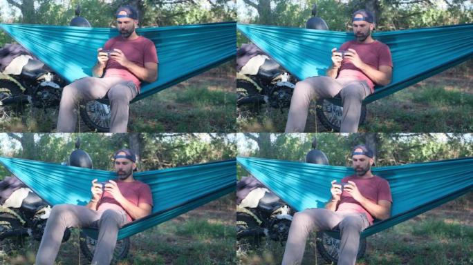 戴着帽子的胡须男子坐在摩托车附近森林的吊床上，看着智能手机上的东西