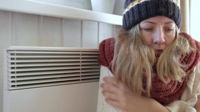 年轻的冷冻女人穿着一件毛衣在冬天的寒冷中颤抖着，脸上表情休克。女孩正在用电加热器呼吸。呆在家里不舒服