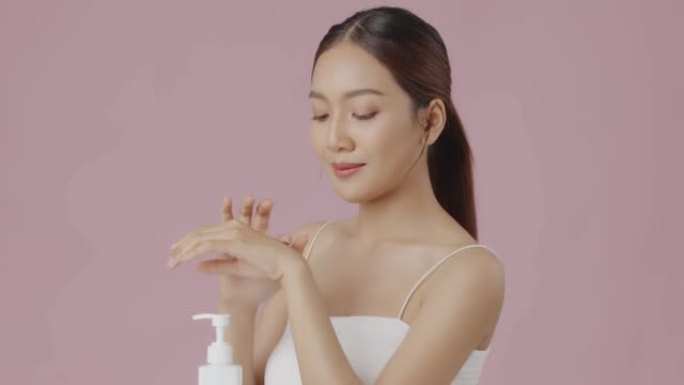 慢动作美丽的年轻亚洲女性在孤立的粉红色背景的手上涂抹保湿面霜乳液。
