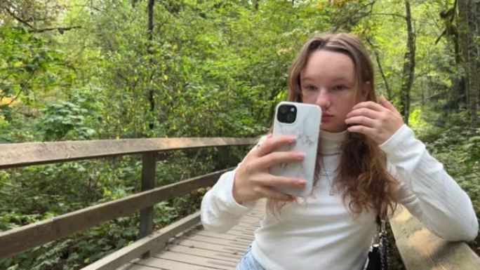 一个穿着白色高领毛衣的女孩拿着手机在她面前自拍，一个白色表壳的女孩少年在大自然的公园里夏天在萨里河上