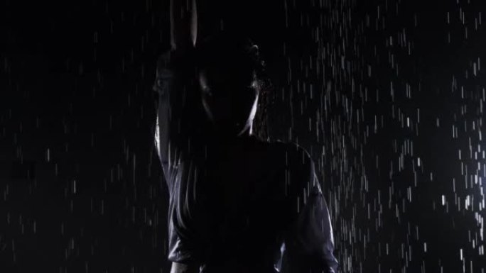 穿着黑色泳衣和衬衫的美丽女孩站在雨中，享受着水滴。黑色背景上的水飞溅。水上工作室。慢动作