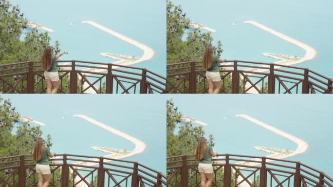 一个女孩站在山顶的栏杆上。她把手指在海里的某个地方。夏季散步。
