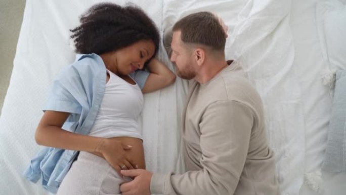 顶视图怀孕的多种族夫妇躺在床上，一起等待分娩。丈夫对女友的温柔照顾。健康怀孕概念