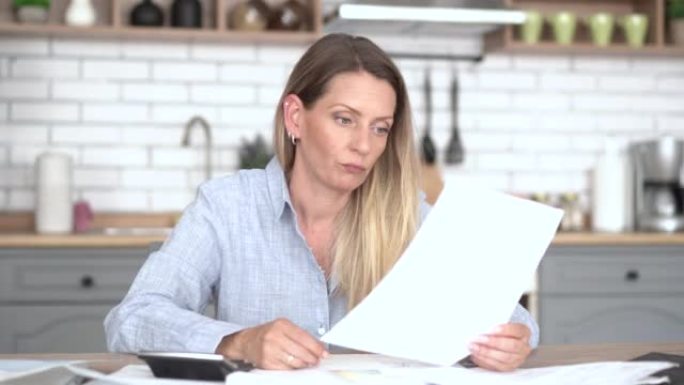 4k视频，一个成熟的女人坐在她公寓的厨房里，计算她的所得税和账单。妇女在经济通货膨胀期间在家中计算抵