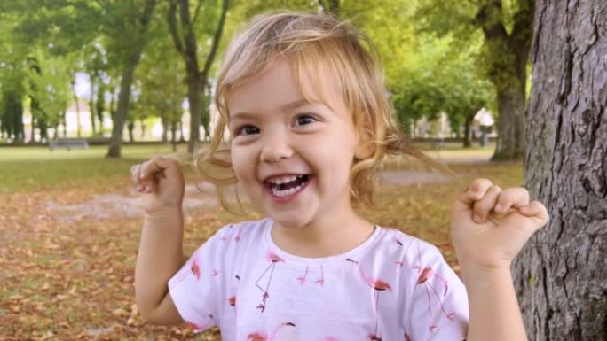 夏天站在公园里看着相机的小女孩。小女孩一边看着镜头一边笑。脸和眼睛有趣的孩子。