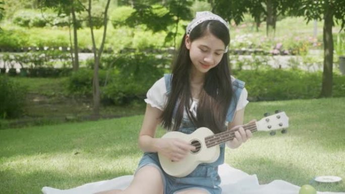 快乐年轻的亚洲女人坐着玩夏威夷四弦琴，放松娱乐是夏天在公园里的爱好，女孩是音乐家，带着积极的生活方式