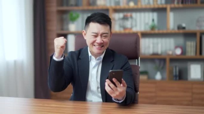 快乐成熟的亚洲商人在办公室用情感看着手机赢家或赢家，金融股体育博彩。男性高兴地惊呼玩游戏。