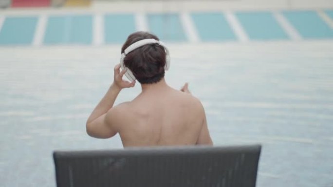 英俊健康的亚洲男性穿着泳衣和耳机听音乐，坐在豪华酒店游泳池旁的床垫上放松，享受休闲活动、生活方式和暑