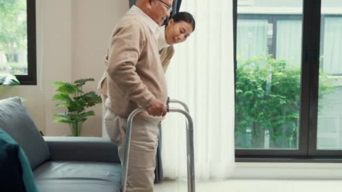 年轻的亚洲女性照顾者教老年男性走路使用助行器在家里的客厅里牵着老年患者的手。