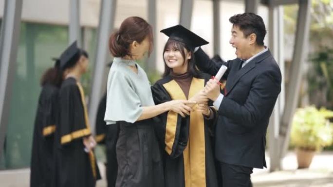 自豪的亚洲华裔父母带着女儿在大学大楼外的大学毕业礼服
