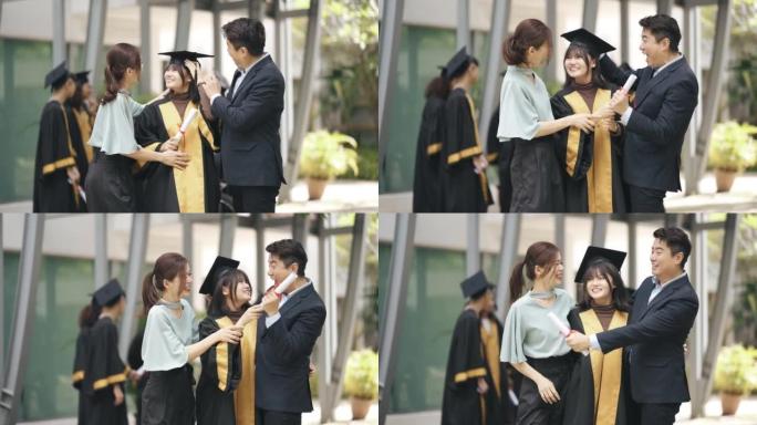 自豪的亚洲华裔父母带着女儿在大学大楼外的大学毕业礼服