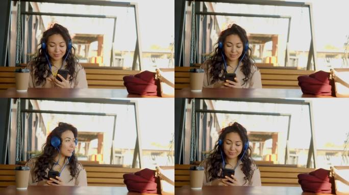 一位戴着耳机的年轻亚洲女性通过智能手机应用程序听音乐，并在社交网络中与朋友积极交流，同时坐在人行道咖