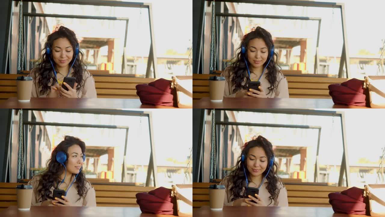 一位戴着耳机的年轻亚洲女性通过智能手机应用程序听音乐，并在社交网络中与朋友积极交流，同时坐在人行道咖