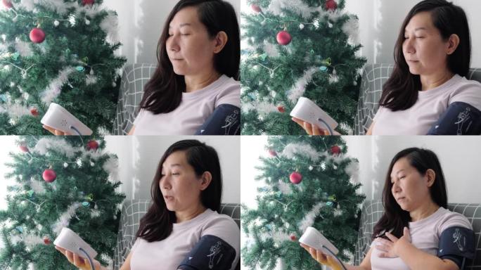 亚洲妇女与血压测试和寻找数字表在网站上的建议，而坐在沙发附近的圣诞树和窗口在家里，健康的生活方式的概