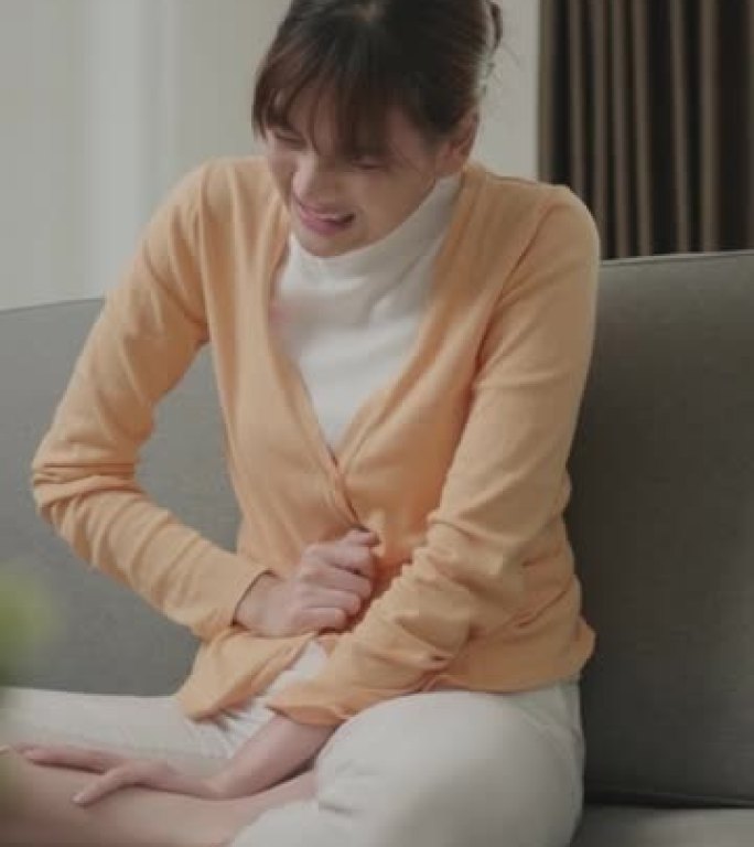 患有严重胃痛的年轻亚洲女性。一个女人坐在沙发上，肚子疼在家里。