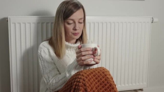 忧心忡忡的女人坐在暖气片附近，在毯子下喝着茶