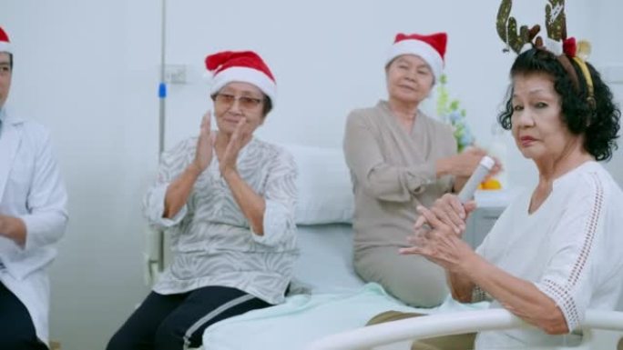 一群戴着圣诞帽的微笑的资深朋友正在唱圣诞歌曲，并与迷人的亚洲医生男性一起拍手。老年男女在养老院的圣诞