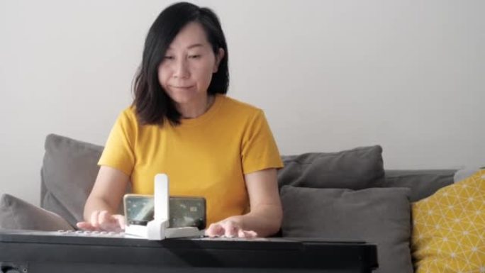 亚洲成年妈妈在家靠窗的沙发上通过移动应用学习弹奏键盘乐器，生活方式概念。