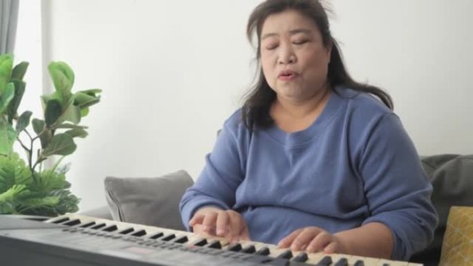 肥胖的亚洲成年妈妈在家学习在靠近窗户的沙发上弹奏键盘乐器，生活方式理念。