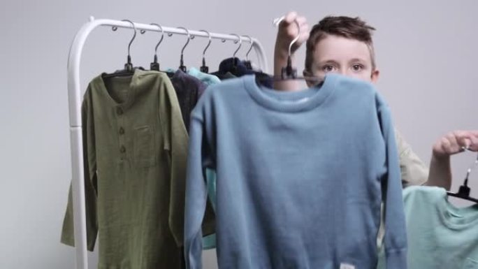 一个时髦的学龄前小男孩在衣架上，拿着一件运动衫和一件t恤向他们展示。