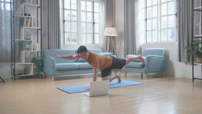亚洲男子在垫子上看着笔记本电脑，在家里摆平衡桌做瑜伽