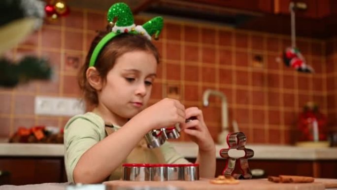 可爱的热情的小女孩穿着厨师围裙和精灵箍，在厨房玩饼干切割机，享受圣诞节的气氛。