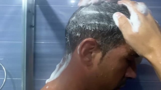 一个男人在浴室里揉着他的头发，用泡沫洗发露在上面起泡沫。一名年轻男子在淋浴时用泡沫抹头发的视频。