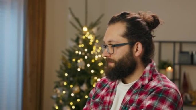 一个留着胡须和眼镜的年轻人站在客厅里。这家伙在找朋友。当他找到他们时，微笑出现在他的脸上。圣诞树上的
