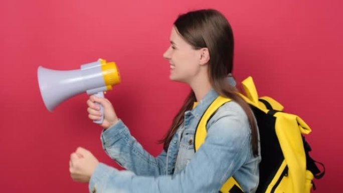 穿着牛仔夹克和黄色背包的肖像年轻女子学生拿着尖叫扩音器宣布折扣销售快点孤立在工作室的红色背景墙上。大