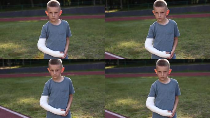 夏天，微笑的断手小男孩坐在户外运动场上。9岁的孩子，四肢骨折，显示另一只健康的手，拇指向上。手臂上的
