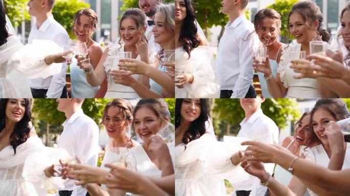 新娘喝香槟，与兴奋的朋友碰杯，欢呼。穿着相同礼服的伴郎和漂亮的伴娘在公园的婚礼上玩得很开心。慢动作。