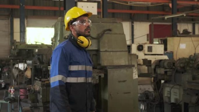 工业工程师回头看着戴着安全镜和带有工厂背景的黄色头盔的相机。
