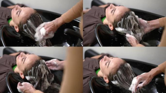 无法辨认的美容师在美容院洗发黑发年轻女子的头发。高加索专业理发师在室内洗女性客户的长发。护发概念。