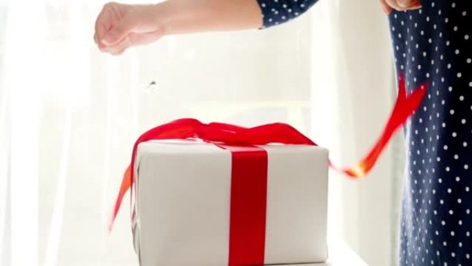 双手从白色礼品盒中拉出红丝带，窗户附近有阳光，假日生活方式概念。