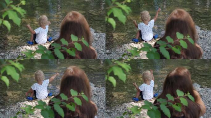 妈妈看着她的小孩在夏天公园的河岸上玩耍，1.5岁的小孩喜欢把鹅卵石扔进水里。