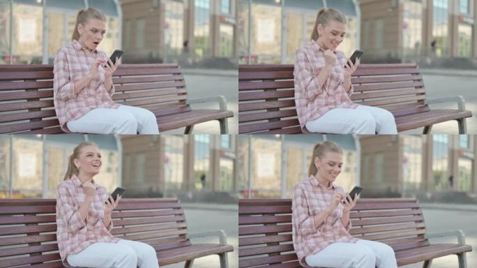 年轻女子坐在户外长凳上庆祝智能手机在线成功