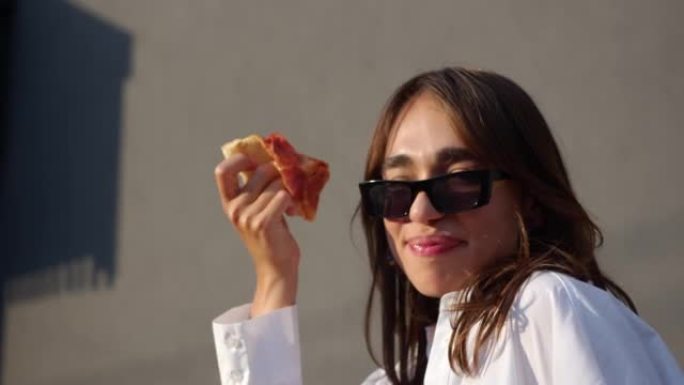 戴墨镜的女孩正在吃披萨。女孩咬披萨，微笑和跳舞