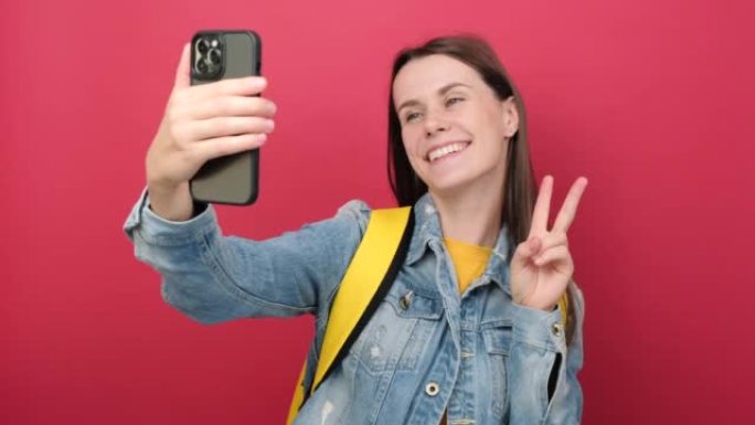 穿着牛仔夹克和黄色背包的年轻女学生的肖像自拍手机，孤立在纯红色背景墙上。高中教育大学学院理念