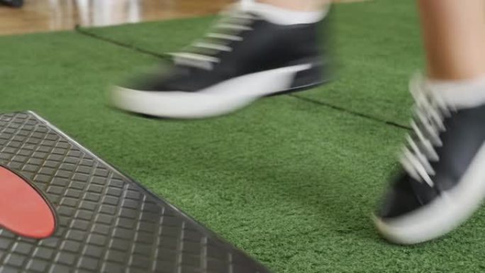 穿着运动鞋的女性腿依次跳上台阶。在带有绿色地板地毯的体育馆中进行有氧运动课程