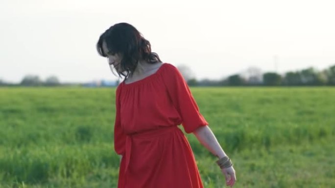 日落或黎明时，穿着红色连衣裙的美丽年轻女子走在绿色的麦田里