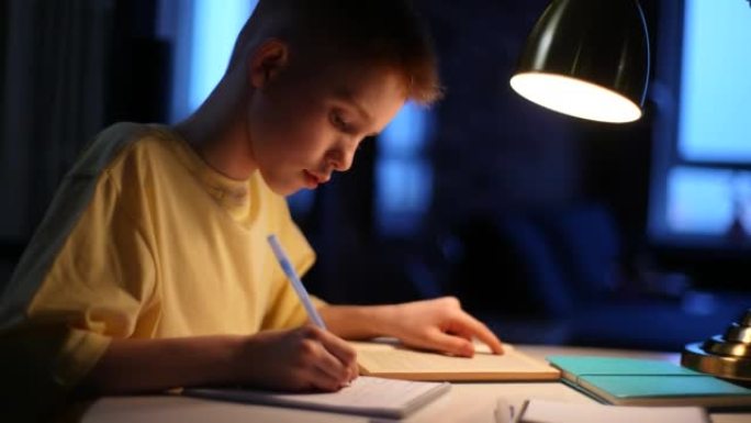 红发男生小学生在家学习的特写侧视图，坐在灯下的桌子旁。